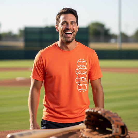 A man smiling in front of a baseball diamond wearing an orange short-sleeve t-shirt, with a colorful graphic of a half baseball with a white graphic of 6 sketched baseballs overlapping, printed on the front of the shirt