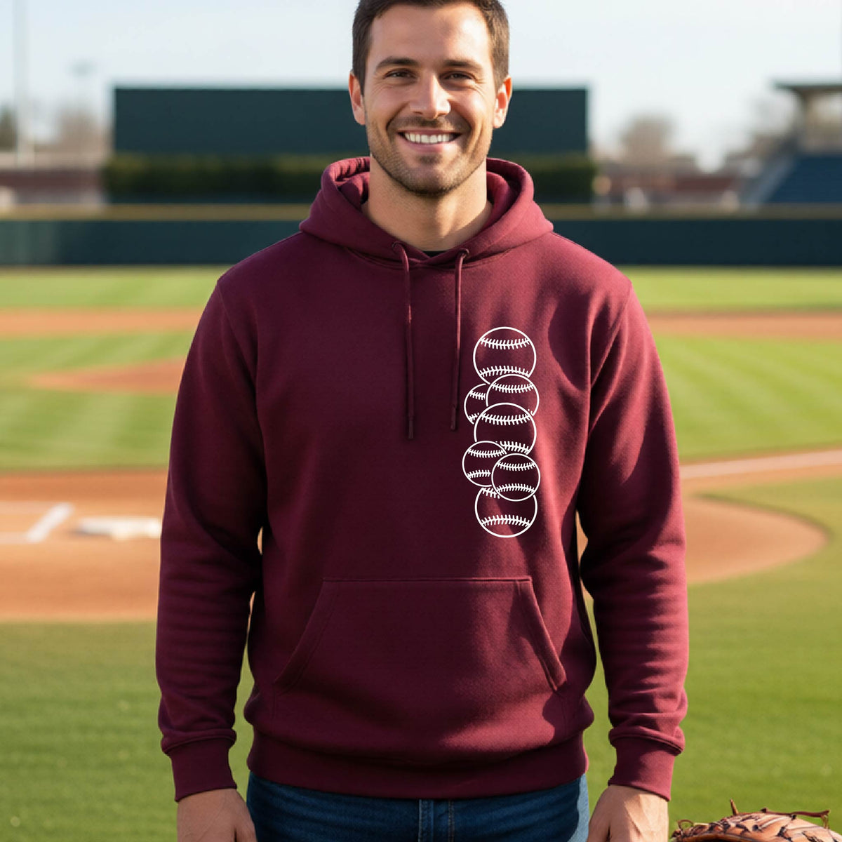 A man smiling in front of a baseball diamond wearing a maroon hoodie pullover, with a white graphic of 6 sketched baseballs overlapping, printed on the front of the shirt