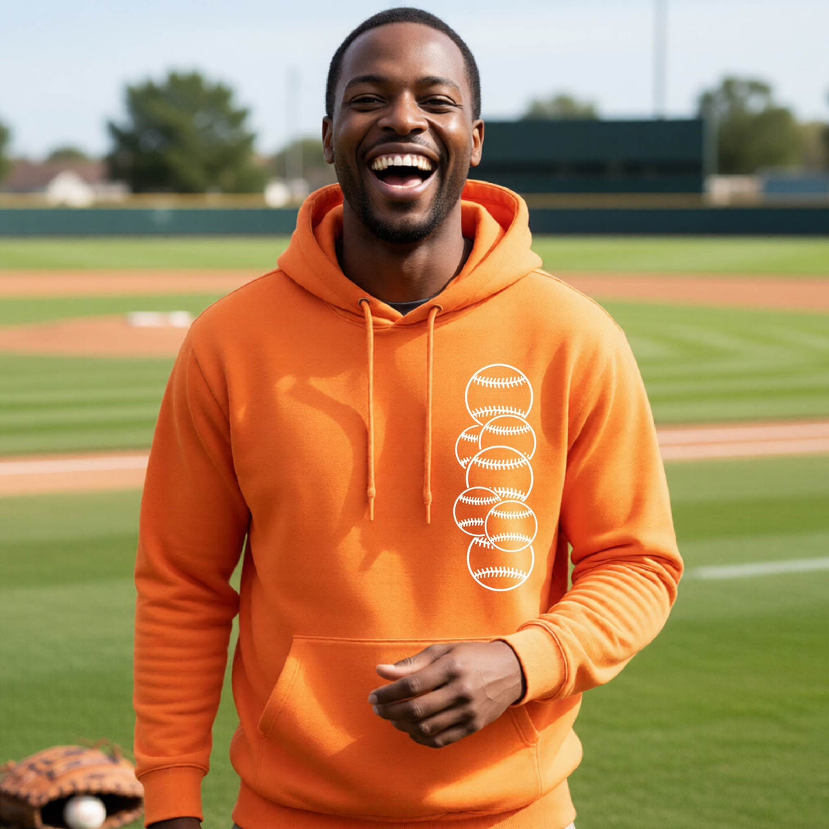 A man smiling in front of a baseball diamond wearing an orange hoodie pullover, with a white graphic of 6 sketched baseballs overlapping, printed on the front of the shirt