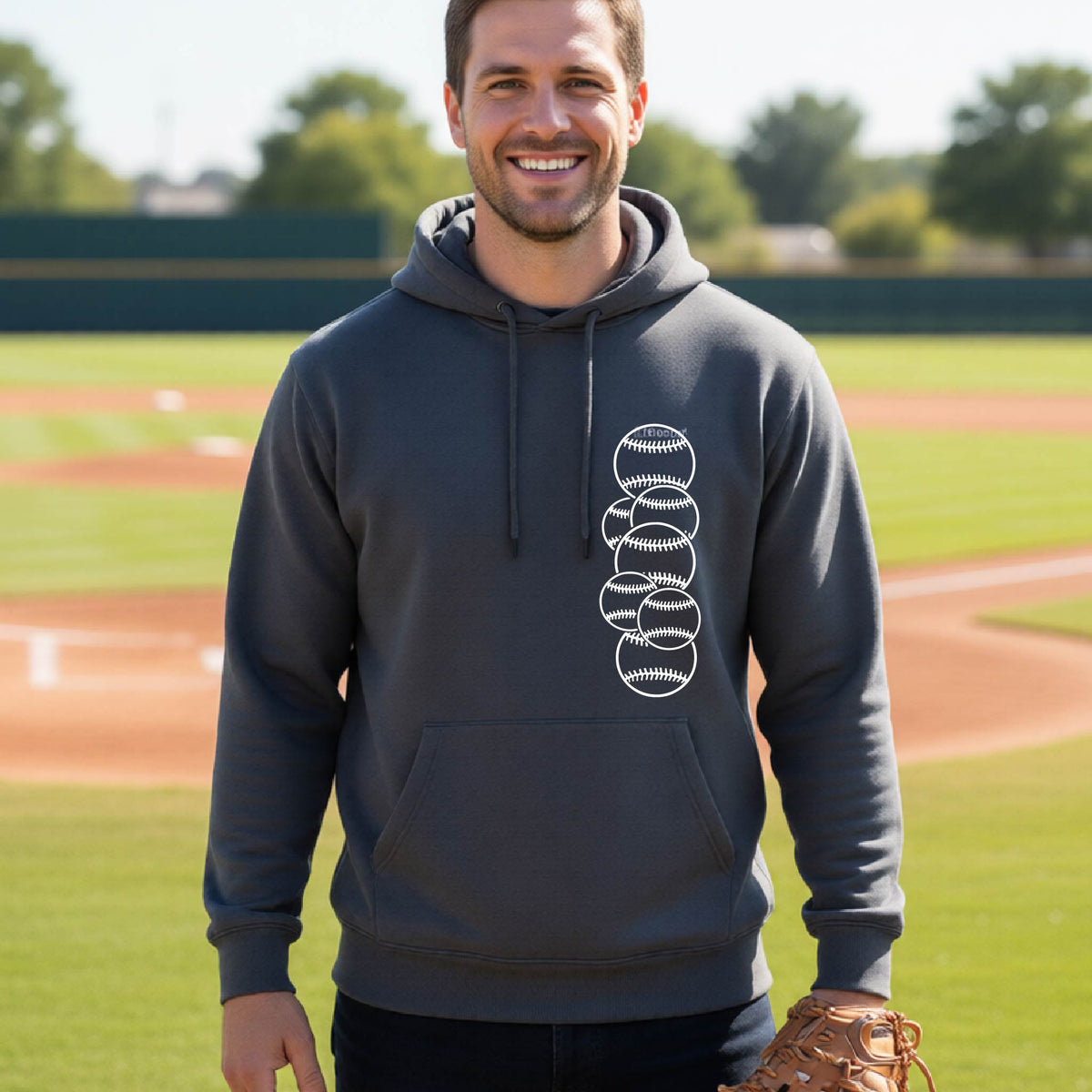 A man smiling in front of a baseball diamond wearing a dark gray hoodie pullover, with a white graphic of 6 sketched baseballs overlapping, printed on the front of the shirt