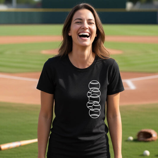 A woman smiling in front of a baseball diamond wearing a black short-sleeve t-shirt, with a colorful graphic of a half baseball with a white graphic of 6 sketched baseballs overlapping, printed on the front of the shirt