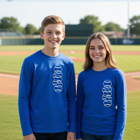 Two youth athletes wearing matching shirts, smiling in front of a baseball diamond wearing royal blue long-sleeve t-shirts, with a white graphic of 6 sketched baseballs overlapping, printed on the front of the shirt.