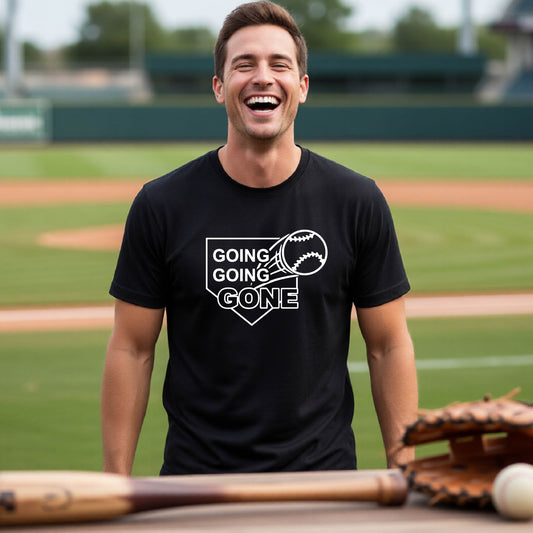 A man smiling in front of a baseball diamond wearing a black short-sleeve t-shirt, with a white graphic of a baseball in flight in front of a home plate with the words ‘GOING GOING GONE’, printed on the front of the shirt.