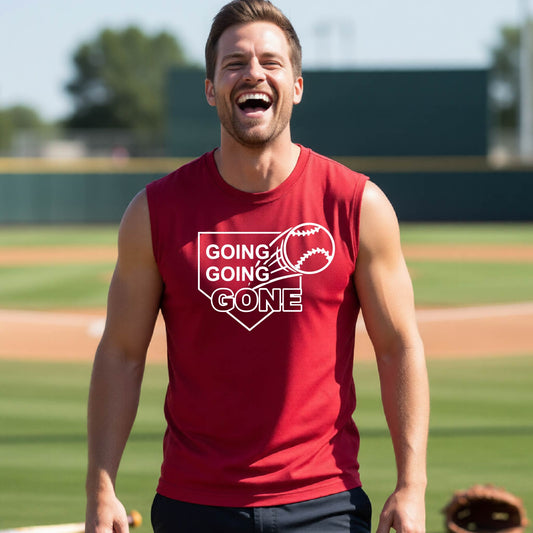 A man smiling in front of a baseball diamond wearing a red sleeveless t-shirt, with a white graphic of a baseball in flight in front of a home plate with the words ‘GOING GOING GONE’, printed on the front of the shirt.