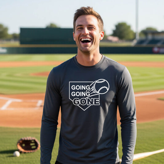 A man smiling in front of a baseball diamond wearing a dark gray long-sleeve t-shirt, with a white graphic of a baseball in flight in front of a home plate with the words ‘GOING GOING GONE’, printed on the front of the shirt.