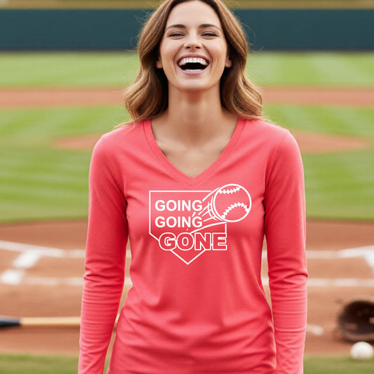 A woman smiling in front of a baseball diamond wearing a coral pink long-sleeve t-shirt, with a white graphic of a baseball in flight in front of a home plate with the words ‘GOING GOING GONE’, printed on the front of the shirt.