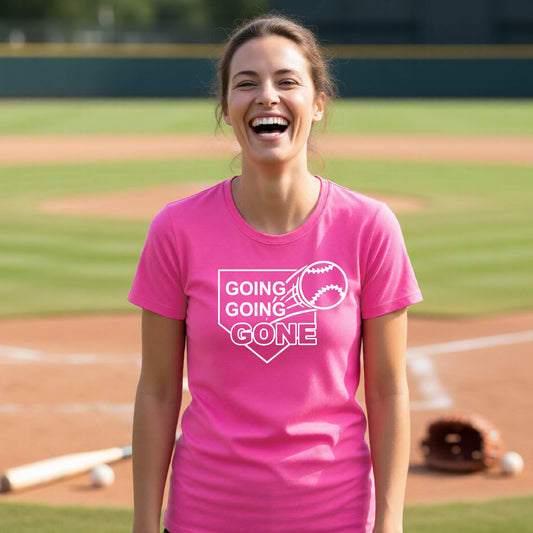 A woman smiling in front of a baseball diamond wearing a pink short-sleeve t-shirt, with a white graphic of a baseball in flight in front of a home plate with the words ‘GOING GOING GONE’, printed on the front of the shirt.