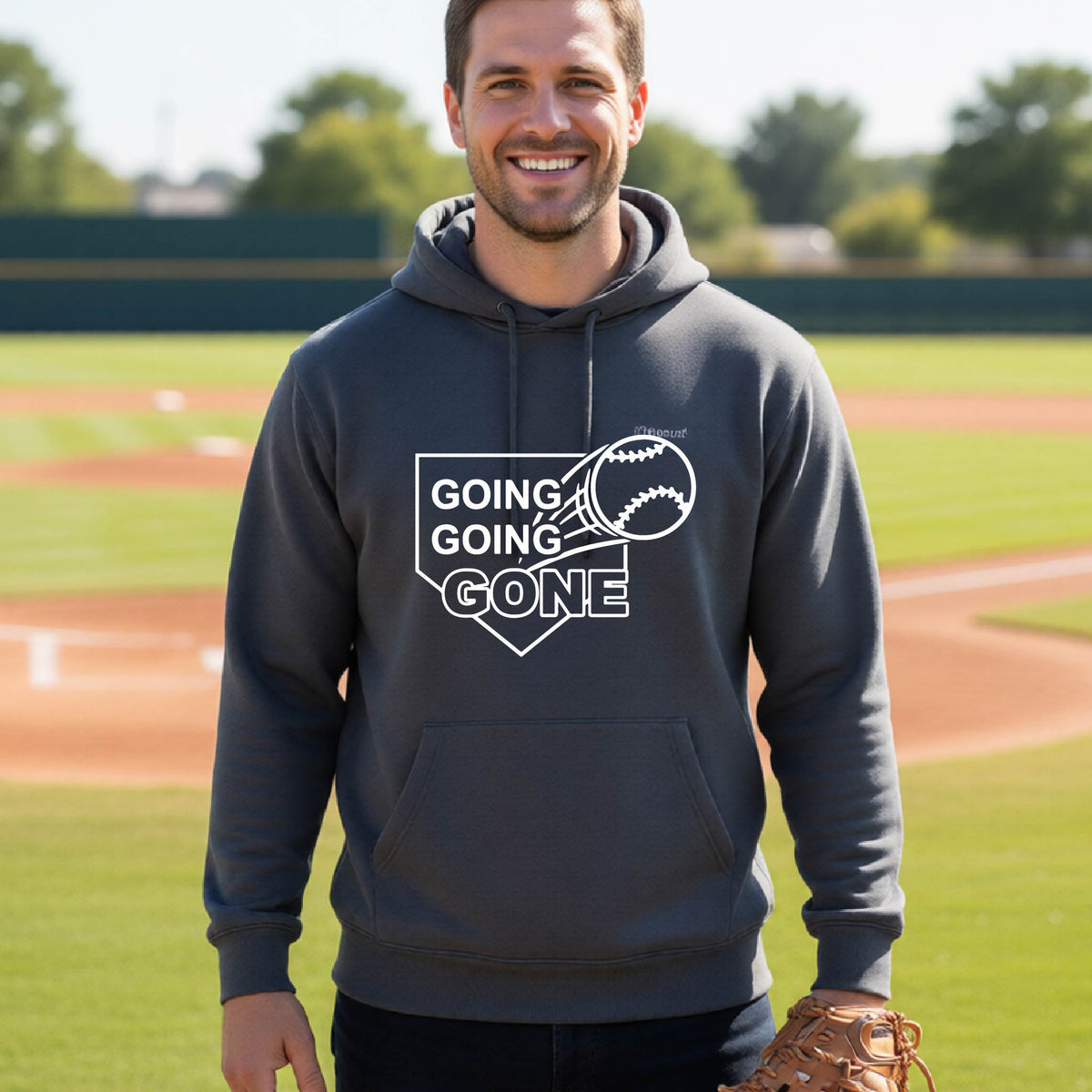 A man smiling in front of a baseball diamond wearing a dark gray hoodie pullover, with a white graphic of a baseball in flight in front of a home plate with the words ‘GOING GOING GONE’, printed on the front of the shirt.