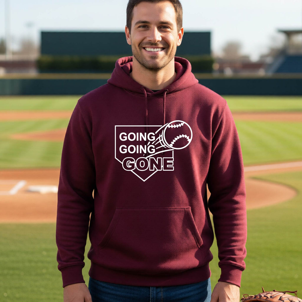 A man smiling in front of a baseball diamond wearing a maroon hoodie pullover, with a white graphic of a baseball in flight in front of a home plate with the words ‘GOING GOING GONE’, printed on the front of the shirt.