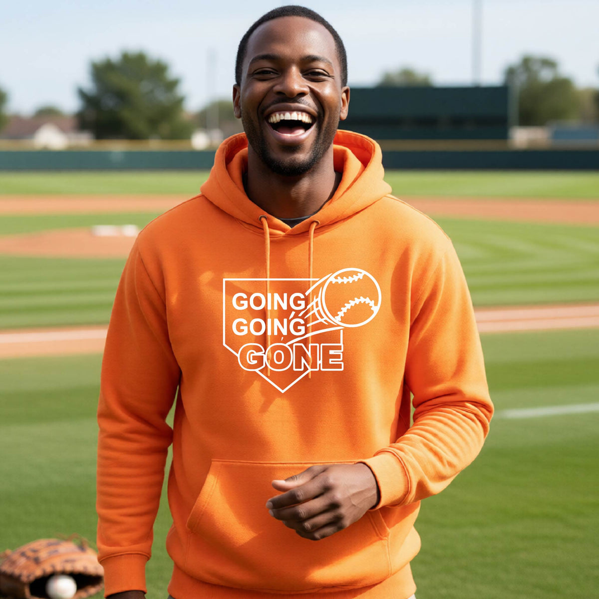 A man smiling in front of a baseball diamond wearing an orange hoodie pullover, with a white graphic of a baseball in flight in front of a home plate with the words ‘GOING GOING GONE’, printed on the front of the shirt.