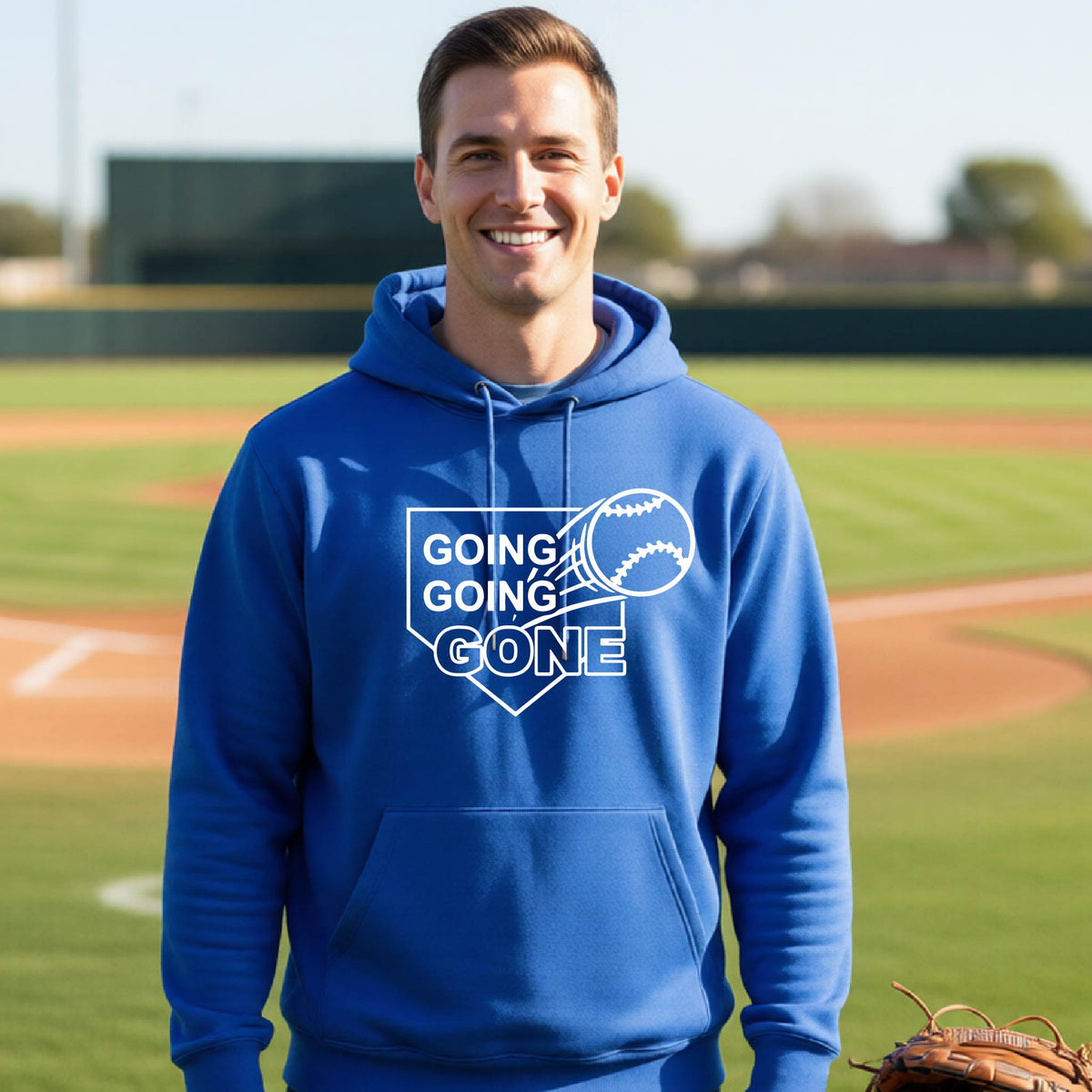 A man smiling in front of a baseball diamond wearing a royal blue hoodie pullover, with a white graphic of a baseball in flight in front of a home plate with the words ‘GOING GOING GONE’, printed on the front of the shirt.