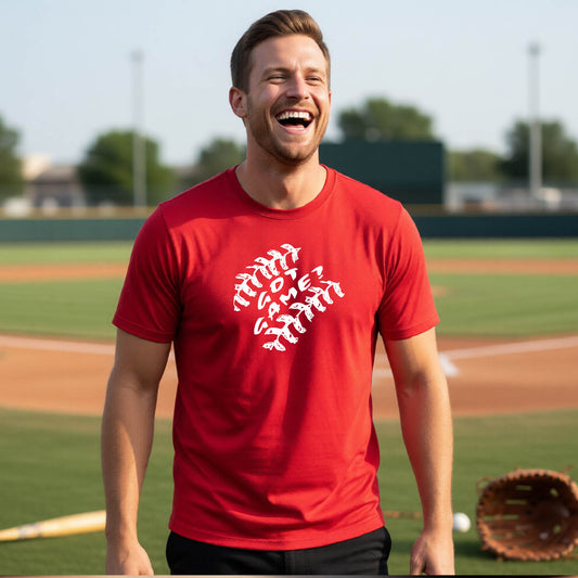 A man smiling in front of a baseball diamond wearing a red short-sleeve t-shirt, with a white graphic of baseball stitches and the words ‘GOT GAME’ inside, printed on the front of the shirt.