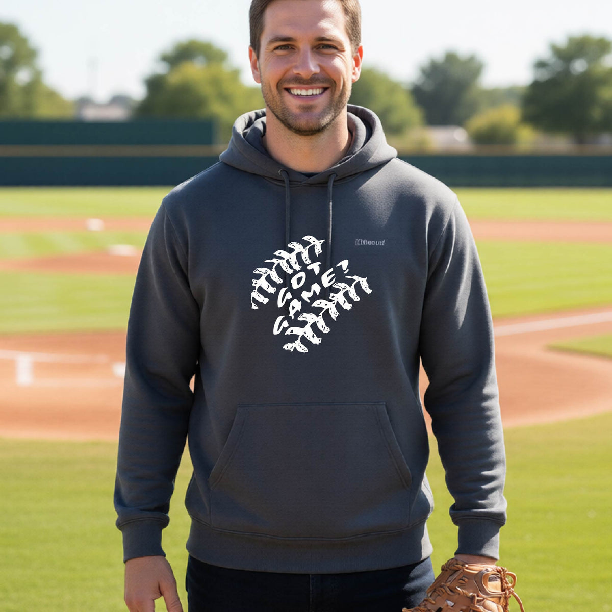 A man smiling in front of a baseball diamond wearing a dark gray pullover hoodie, with a white graphic of baseball stitches and the words ‘GOT GAME’ inside, printed on the front of the shirt.