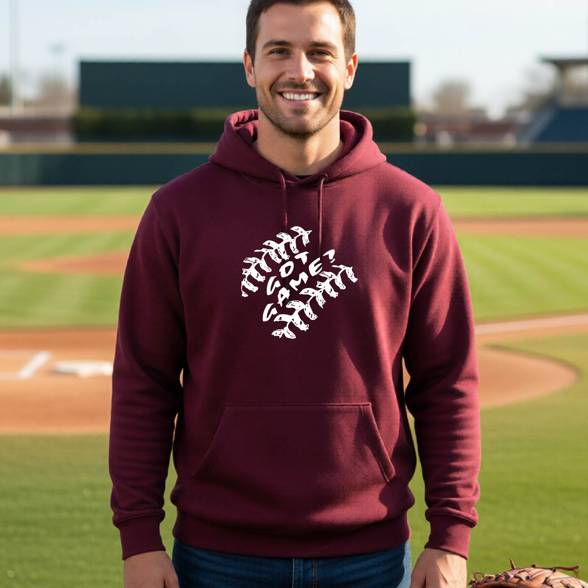A man smiling in front of a baseball diamond wearing a maroon pullover hoodie, with a white graphic of baseball stitches and the words ‘GOT GAME’ inside, printed on the front of the shirt.