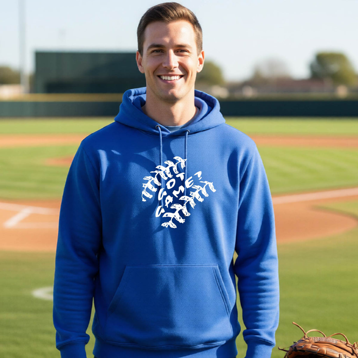 A man smiling in front of a baseball diamond wearing a royal blue pullover hoodie, with a white graphic of baseball stitches and the words ‘GOT GAME’ inside, printed on the front of the shirt.