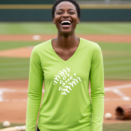 A woman smiling in front of a baseball diamond wearing a lime green long-sleeve t-shirt, with a white graphic of baseball stitches and the words ‘GOT GAME’ inside, printed on the front of the shirt.