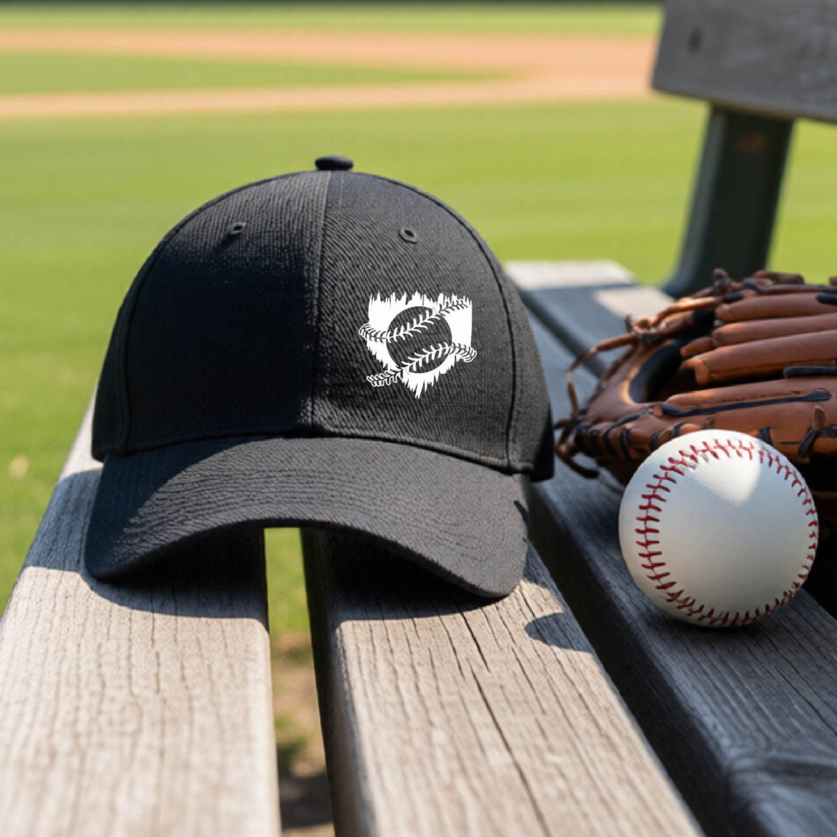 A close-up of a black baseball hat on an outdoor bench in front of a baseball diamond, with an artistic white graphic of a sketch of baseball home plate with baseball stitches across the front, printed on the front of the shirt.