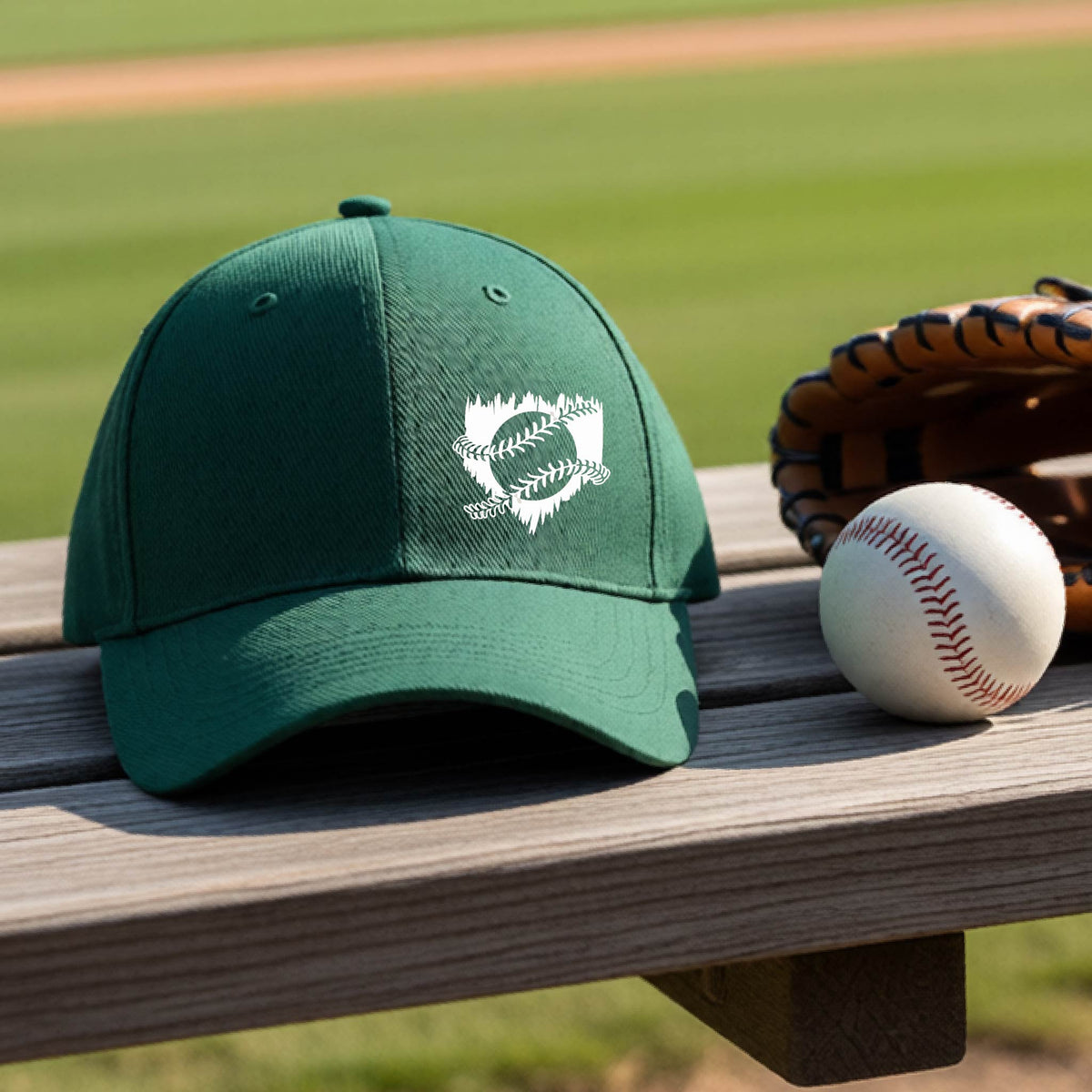 A close-up of a green baseball hat on an outdoor bench in front of a baseball diamond, with an artistic white graphic of a sketch of baseball home plate with baseball stitches across the front, printed on the front of the shirt.