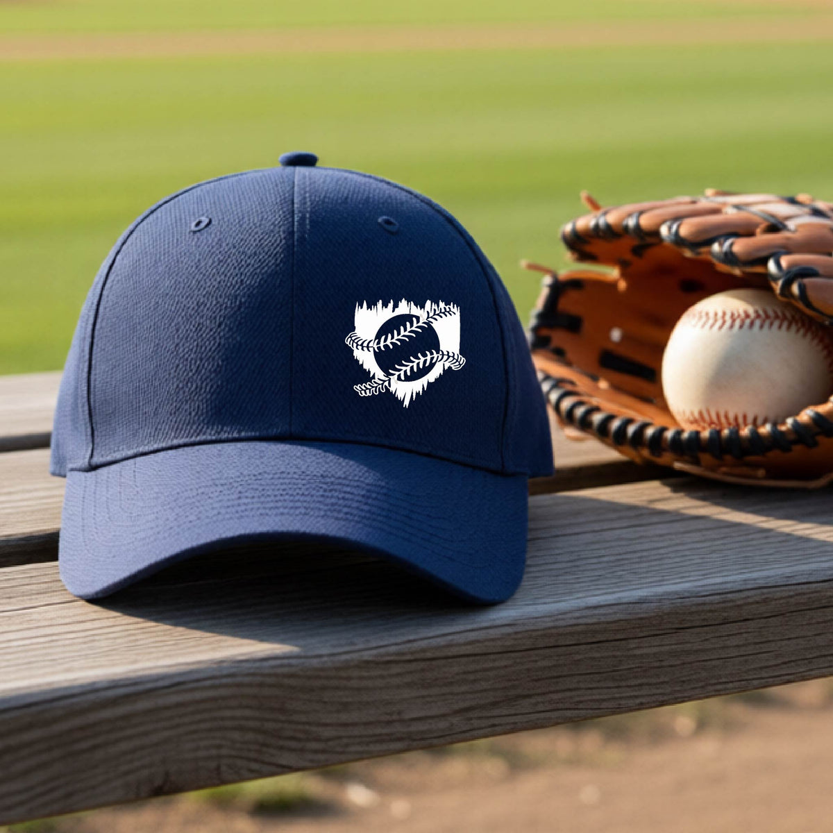 A close-up of a navy blue baseball hat on an outdoor bench in front of a baseball diamond, with an artistic white graphic of a sketch of baseball home plate with baseball stitches across the front, printed on the front of the shirt.