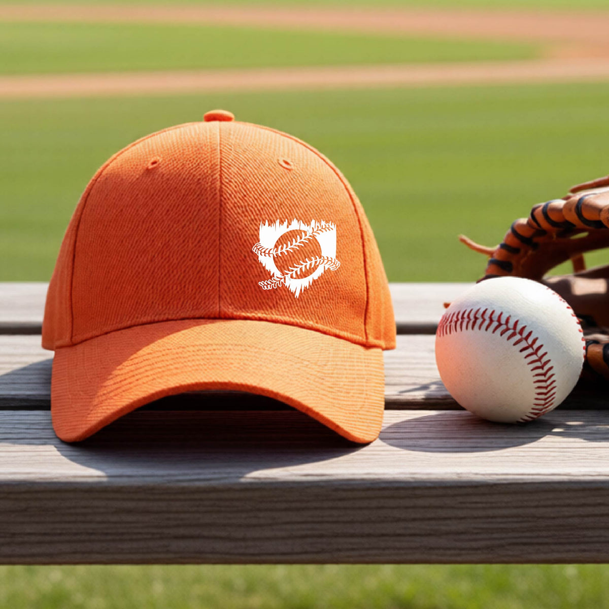 A close-up of an orange baseball hat on an outdoor bench in front of a baseball diamond, with an artistic white graphic of a sketch of baseball home plate with baseball stitches across the front, printed on the front of the shirt.