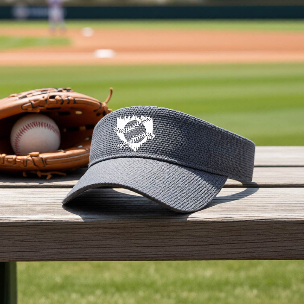 A close-up of a graphite gray baseball visor on an outdoor bench in front of a baseball diamond, with an artistic white graphic of a sketch of baseball home plate with baseball stitches across the front, printed on the front of the shirt.