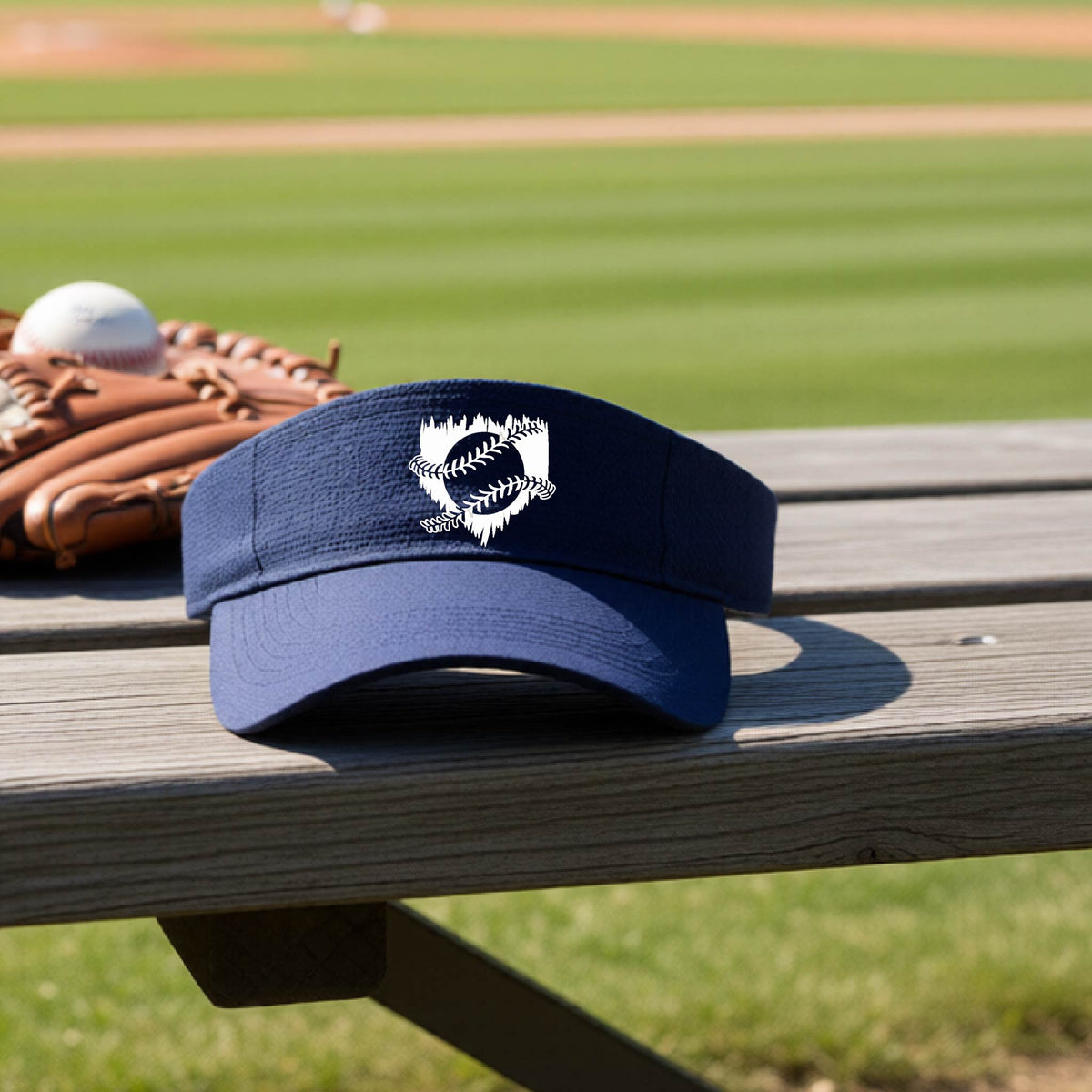 A close-up of a navy blue baseball visor on an outdoor bench in front of a baseball diamond, with an artistic white graphic of a sketch of baseball home plate with baseball stitches across the front, printed on the front of the shirt.