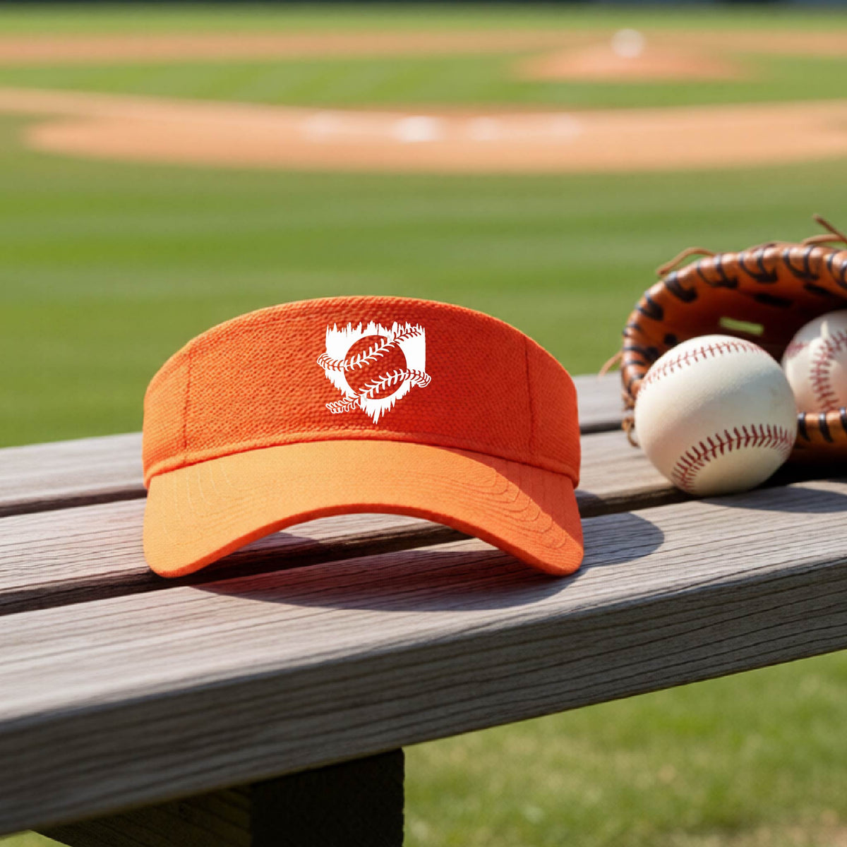 A close-up of an orange baseball visor on an outdoor bench in front of a baseball diamond, with an artistic white graphic of a sketch of baseball home plate with baseball stitches across the front, printed on the front of the shirt.