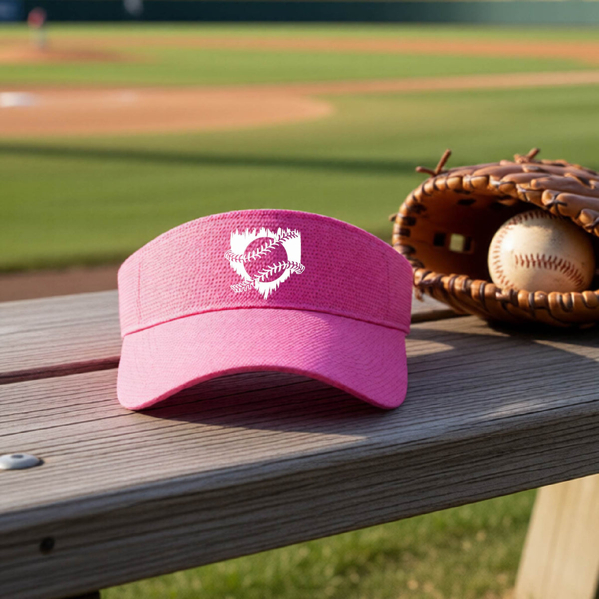 A close-up of a pink baseball visor on an outdoor bench in front of a baseball diamond, with an artistic white graphic of a sketch of baseball home plate with baseball stitches across the front, printed on the front of the shirt.