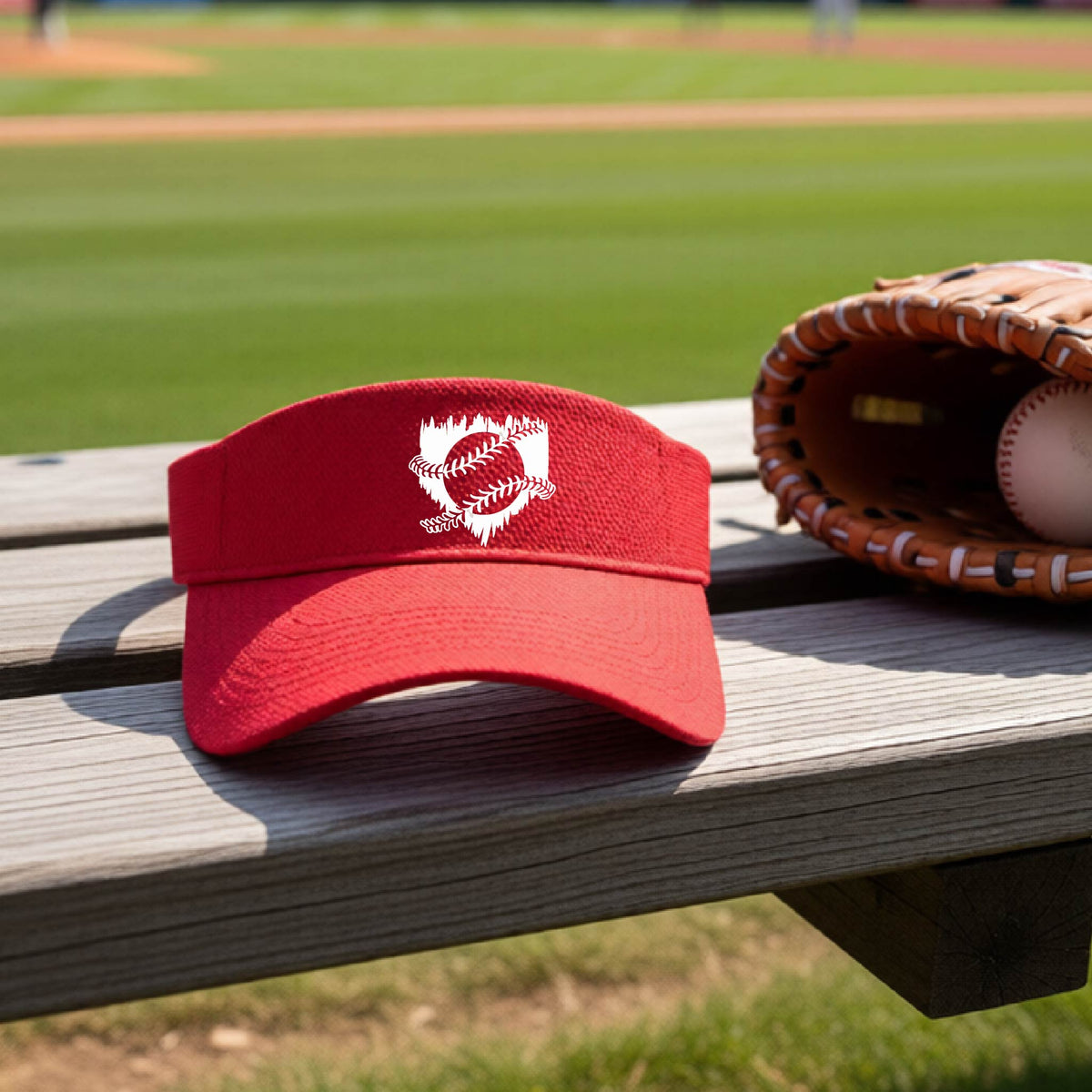 A close-up of a red baseball visor on an outdoor bench in front of a baseball diamond, with an artistic white graphic of a sketch of baseball home plate with baseball stitches across the front, printed on the front of the shirt.