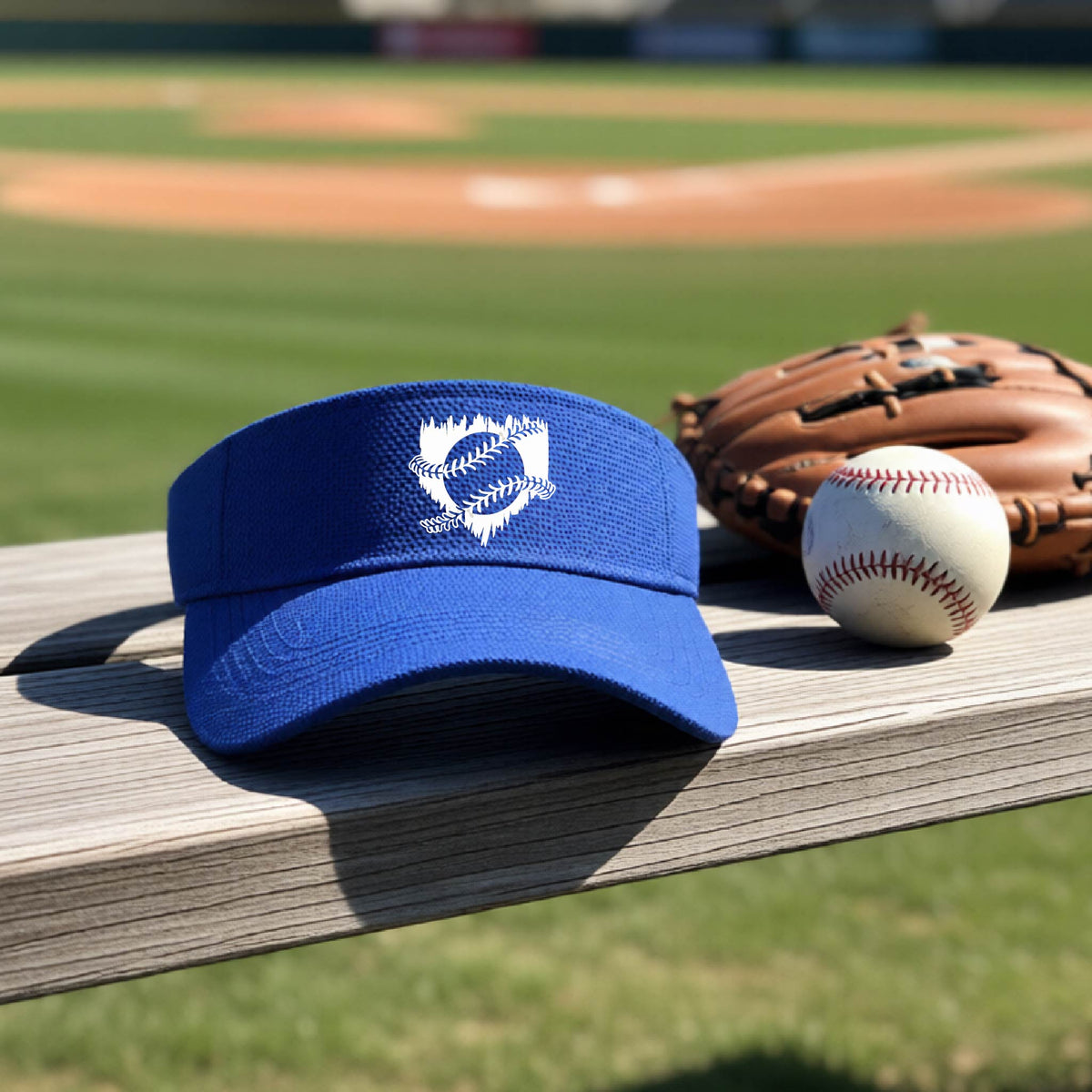 A close-up of a royal blue baseball visor on an outdoor bench in front of a baseball diamond, with an artistic white graphic of a sketch of baseball home plate with baseball stitches across the front, printed on the front of the shirt.