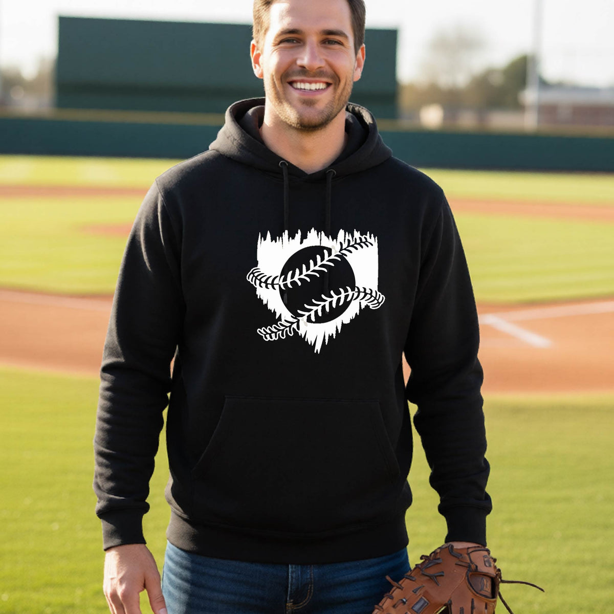 A man smiling in front of a baseball diamond wearing a black pullover hoodie, with an artistic white graphic of a sketch of baseball home plate with baseball stitches across the front, printed on the front of the shirt.