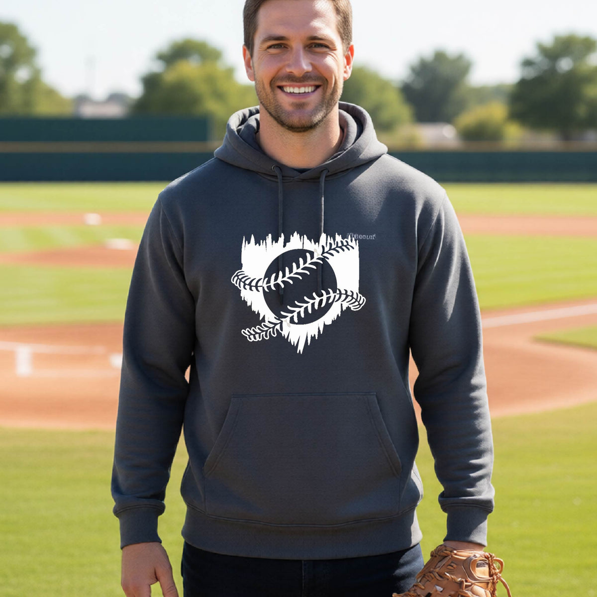 A man smiling in front of a baseball diamond wearing a dark gray pullover hoodie, with an artistic white graphic of a sketch of baseball home plate with baseball stitches across the front, printed on the front of the shirt.