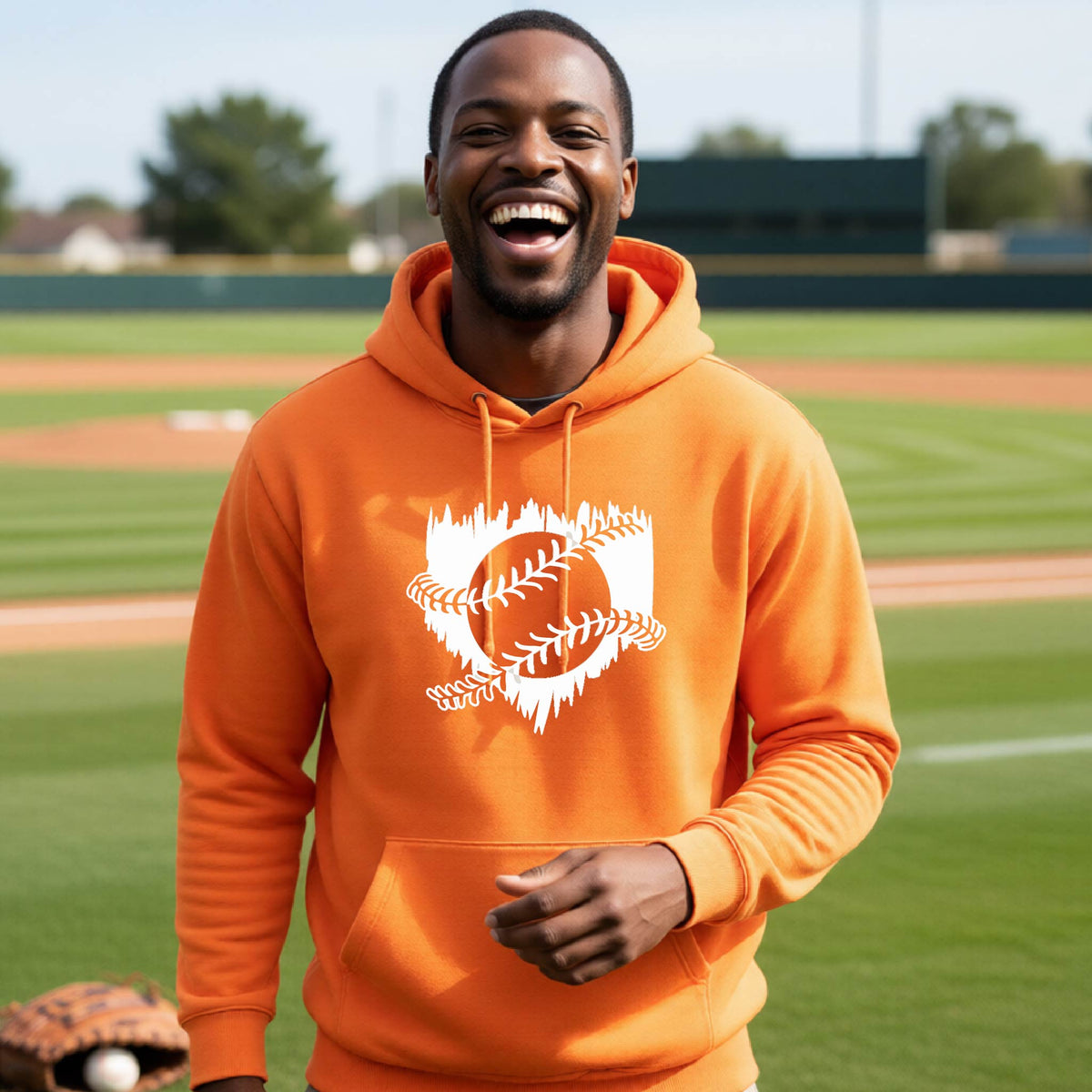 A man smiling in front of a baseball diamond wearing an orange pullover hoodie, with an artistic white graphic of a sketch of baseball home plate with baseball stitches across the front, printed on the front of the shirt.