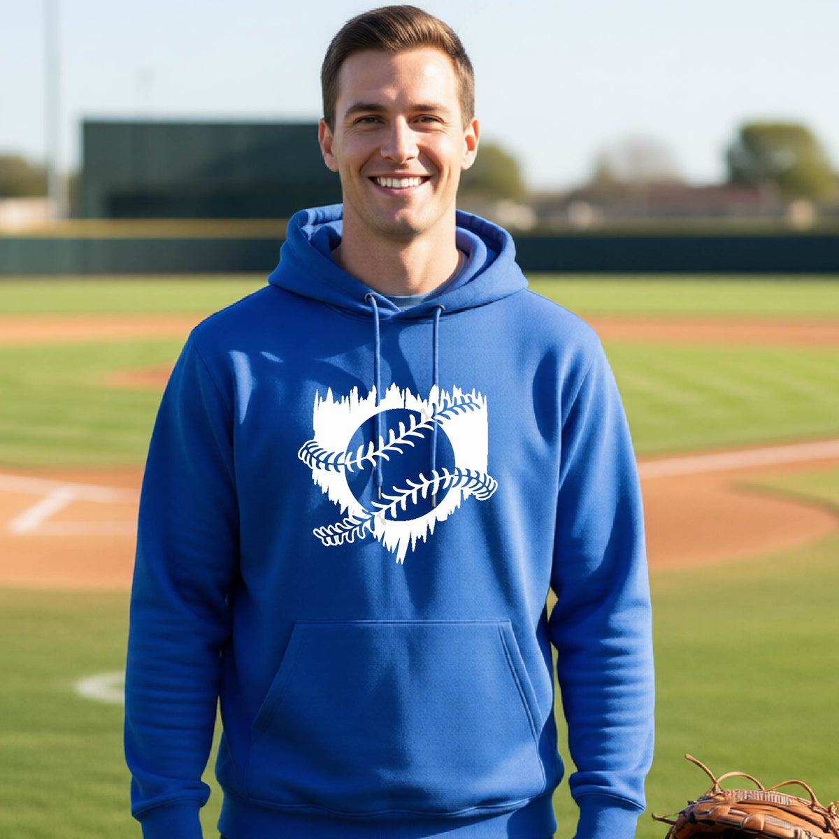 A man smiling in front of a baseball diamond wearing a royal blue pullover hoodie, with an artistic white graphic of a sketch of baseball home plate with baseball stitches across the front, printed on the front of the shirt.