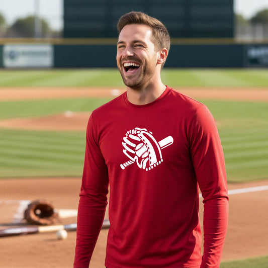 A man smiling in front of a baseball diamond wearing a red long-sleeve t-shirt with a white graphic of baseball itt, bat and ball, printed on the front.