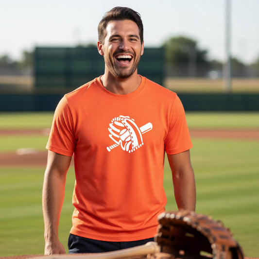 A man smiling in front of a baseball diamond wearing an orange short-sleeve t-shirt with a white graphic of baseball mitt, bat and ball, printed on the front.