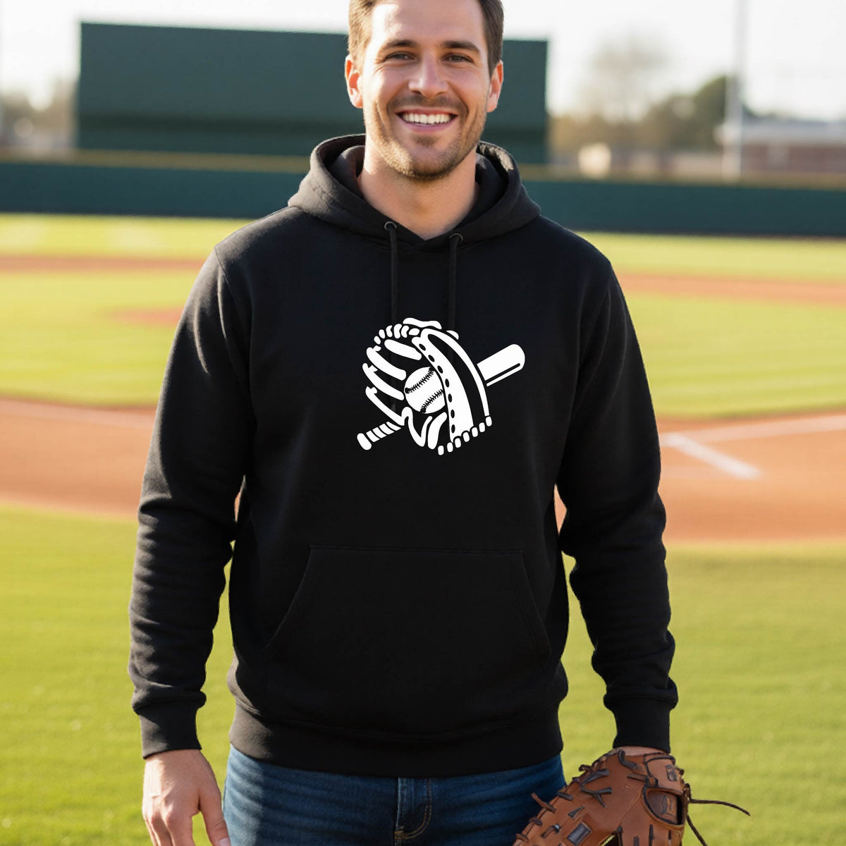 A man smiling in front of a baseball diamond wearing a black hoodie pullover with a white graphic of baseball itt, bat and ball, printed on the front.