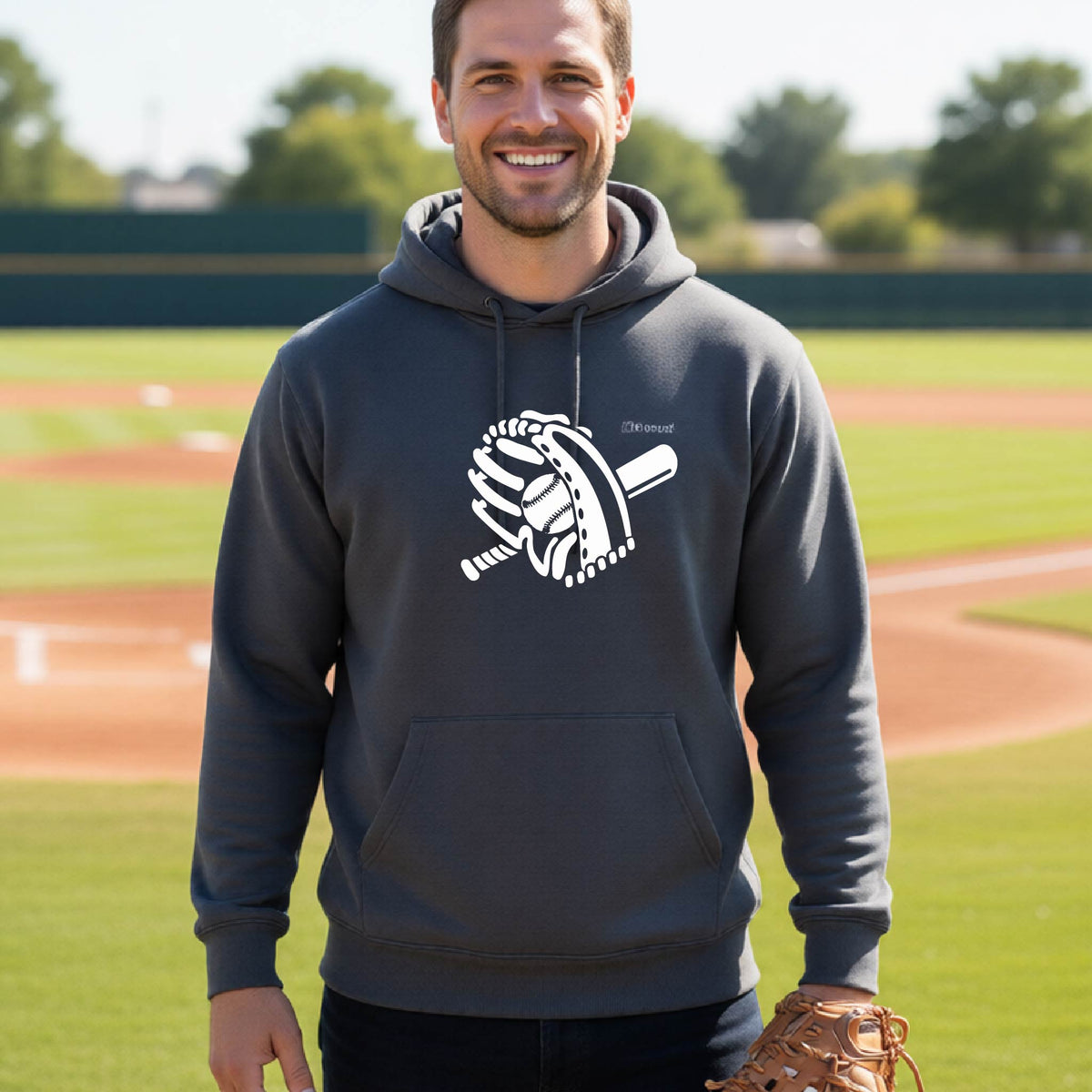 A man smiling in front of a baseball diamond wearing a dark gray hoodie pullover with a white graphic of baseball itt, bat and ball, printed on the front.