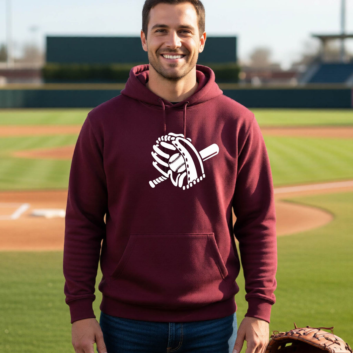 A man smiling in front of a baseball diamond wearing a maroon hoodie pullover with a white graphic of baseball itt, bat and ball, printed on the front.