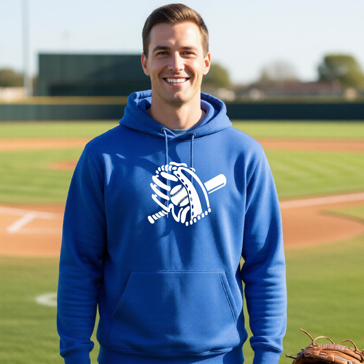A man smiling in front of a baseball diamond wearing a royal blue hoodie pullover with a white graphic of baseball itt, bat and ball, printed on the front.