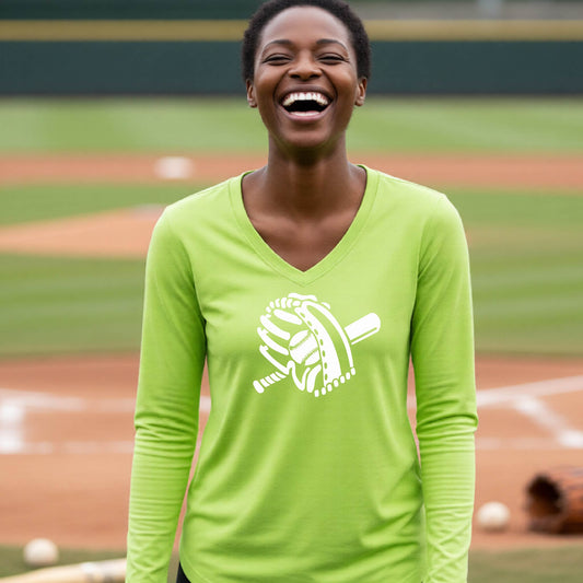 A woman smiling in front of a baseball diamond wearing a lime green long-sleeve t-shirt with a white graphic of baseball mitt, bat and ball, printed on the front.