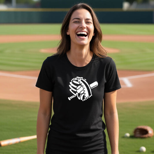 A woman smiling in front of a baseball diamond wearing a black short-sleeve t-shirt with a white graphic of baseball itt, bat and ball, printed on the front.