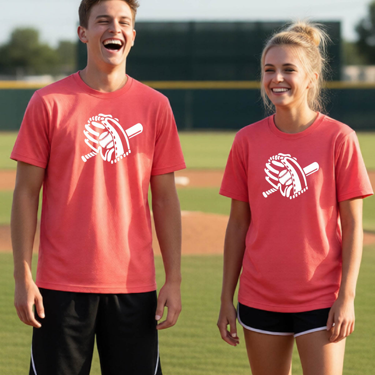 Two young athletes wearing matching shirts, smiling in front of a baseball diamond wearing coral colored short-sleeve t-shirts with a white graphic of baseball mitt, bat and ball, printed on the front.
