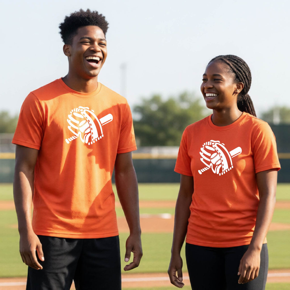 Two young athletes wearing matching shirts, smiling in front of a baseball diamond wearing orange colored short-sleeve t-shirts with a white graphic of baseball mitt, bat and ball, printed on the front.