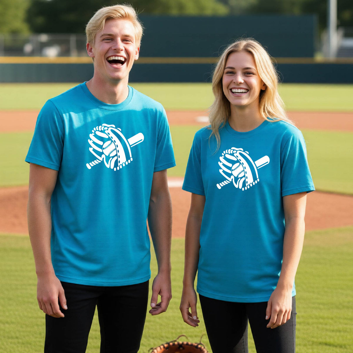 Two young athletes wearing matching shirts, smiling in front of a baseball diamond wearing bright blue colored short-sleeve t-shirts with a white graphic of baseball mitt, bat and ball, printed on the front.  