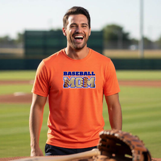 A man smiling in front of a baseball diamond wearing an orange short-sleeve t-shirt with a colorful graphic of the words ‘BASEBALL MOM’ printed on the front.