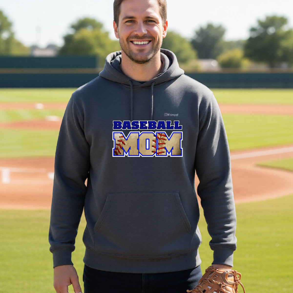 A man smiling in front of a baseball diamond wearing a dark gray hoodie pullover with a colorful graphic of the words ‘BASEBALL MOM’ printed on the front.