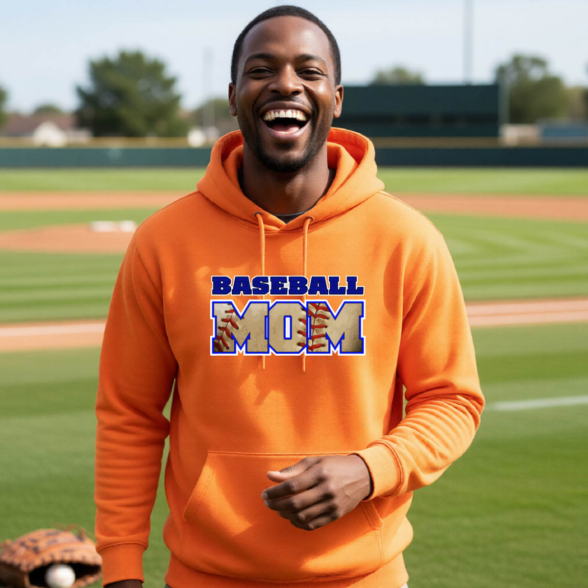 A man smiling in front of a baseball diamond wearing an orange hoodie pullover with a colorful graphic of the words ‘BASEBALL MOM’ printed on the front.