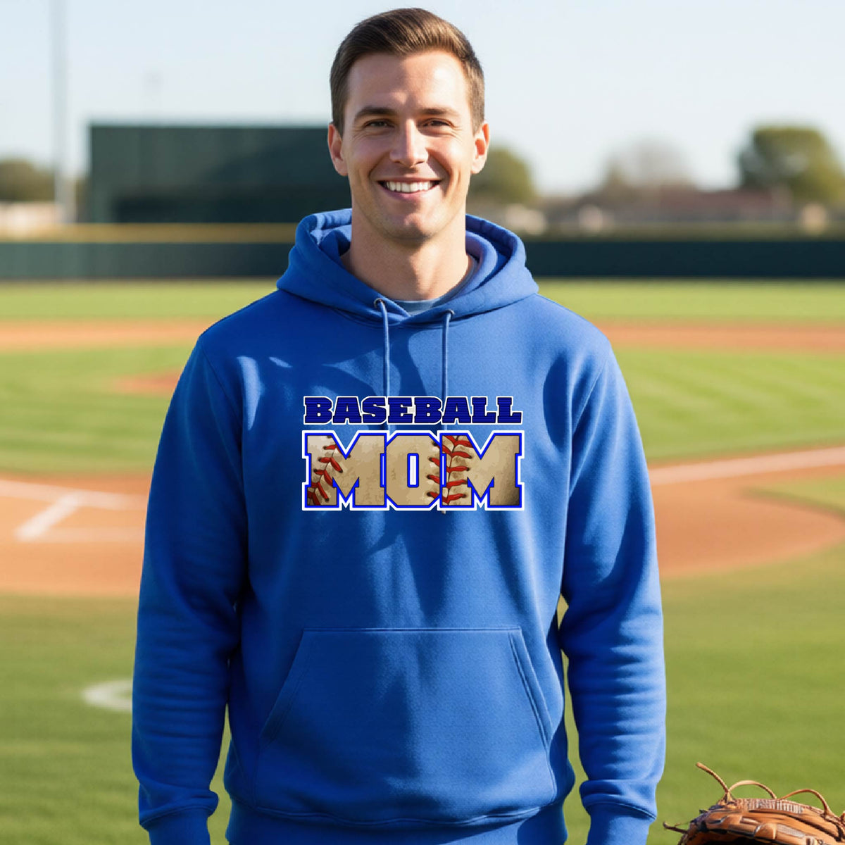 A man smiling in front of a baseball diamond wearing a royal blue hoodie pullover with a colorful graphic of the words ‘BASEBALL MOM’ printed on the front.