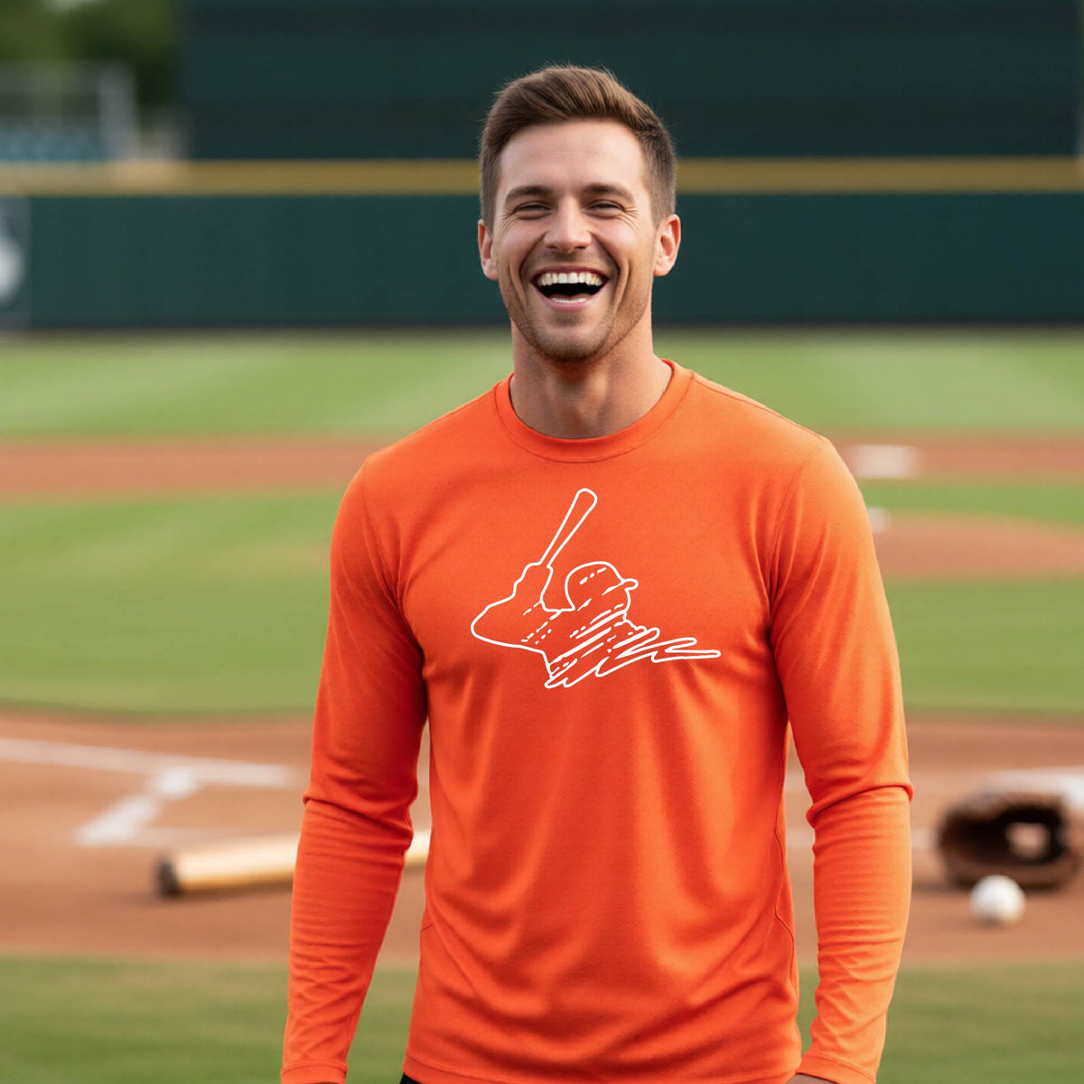 A man smiling in front of a baseball diamond wearing an orange long-sleeve t-shirt, with a white graphic of an outline of a baseball player up to bat, printed on the front.
